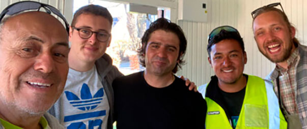 Five men pose for a selfie indoors, two wearing high-visibility vests, smiling at the camera.