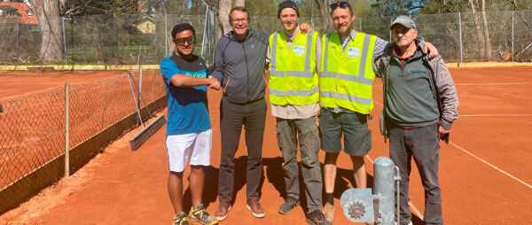 Five men stand on a red clay tennis court near a chain-link fence, two wearing high-visibility vests, posing for the photo with equipment nearby.